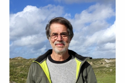 Met duinen en een blauwe lucht op de achtergrond een foto van een man met grijze trui, groene jas, een baard en een bril. De man kijkt recht in de camera